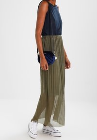 Person som bär en marinblå ärmlös topp, en olivgrön genomskinlig plisserad maxi kjol, vita plattformssneakers och en liten marinblå kviltad crossbody-väska.