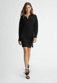 Black shirt dress with a pocket, side detailing of beads, and a slit. Paired with black sandals featuring feather accents. Smooth texture.