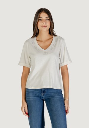 T-shirt beige chiaro con scollo a V, maniche corte e dettagli del logo ricamati. Tessuto morbido e vestibilità comoda, abbinato a jeans blu.