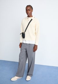 Jersey de color crema con cuello, pantalones anchos grises y zapatillas crema. Bolso cruzado negro con un cierre amarillo.