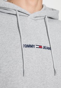 Felpa grigia con cappuccio realizzata in tessuto morbido, con un logo ricamato "TOMMY JEANS" in blu navy, rosso e bianco sul petto.