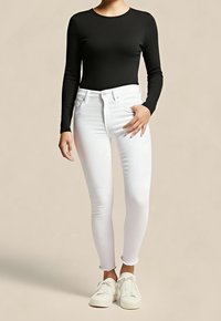 Femme portant un haut noir à manches longues, un jean skinny blanc et des baskets blanches, debout devant un fond beige uni.