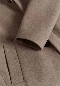 Brauner Wollmantel mit einer weichen, strukturierten Oberfläche, ausgestattet mit einem schwarzen Knopf und einer Seitentasche, die ein klares, maßgeschneidertes Design betont.