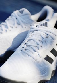 Pár bielych Adidas športových topánok s čiernymi pruhmi a šnúrkami, uložených na tmavom textúrovanom povrchu.