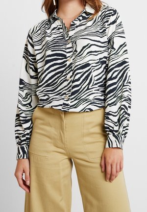 Vrouw draagt een langärmelig zebragestreept blouse met knopen, ingestopt in een beige broek met een hoge taille, met zichtbare gouden kettingen om de nek.