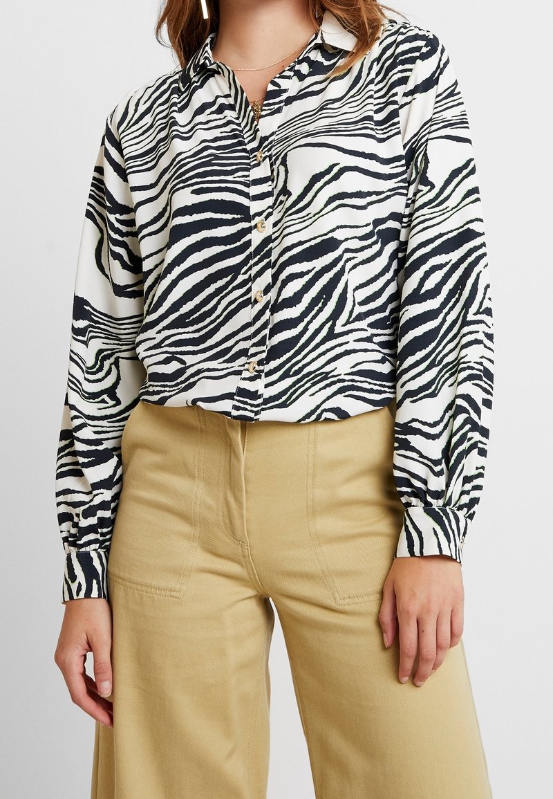 Femme portant une chemise boutonnée à manches longues à rayures zèbre rentrée dans un pantalon beige taille haute, avec des colliers dorés visibles autour du cou.