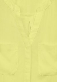 Blusa gialla con scollatura a V, maniche corte, due tasche sul petto e una texture di tessuto pulita e liscia.