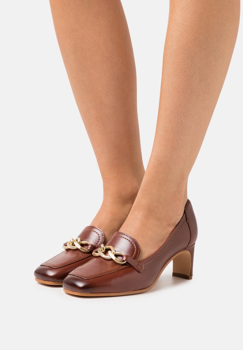 Scarpe con tacco alto in pelle marrone con punta quadrata, caratterizzate da un accento in catena dorata sulla parte anteriore e una finitura liscia e texturizzata.