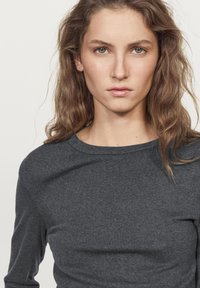 Ciemnoszara koszula z długim rękawem, wykonana z miękkiego materiału, z okrągłym dekoltem i gładką fakturą. Modelka ma średniej długości fale włosów.