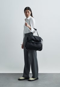 Borsa tote nera imbottita con design elegante e tracolla regolabile, tenuta da una persona con camicia bianca e pantaloni grigi su uno sfondo neutro.