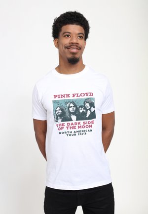 Uomo sorridente, che indossa una maglietta bianca dei Pink Floyd con la scritta "The Dark Side of the Moon North American Tour 1973" e foto dei membri della band.