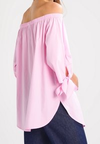 Top in cotone leggero rosa pallido off-the-shoulder con scollo elastico, maniche a sbuffo e dettagli annodati ai polsini. Orlo leggermente curvo.