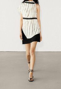 Robe asymétrique à motifs rayés blanc et noir, avec un col montant, un accent noir à la taille, associée à des sandales à lanières noires.