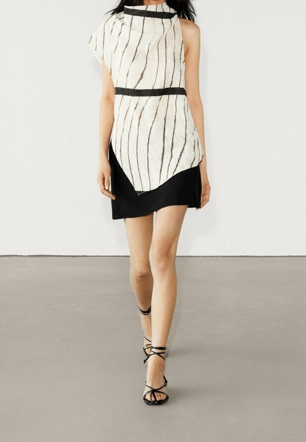 Robe asymétrique à motifs rayés blanc et noir, avec un col montant, un accent noir à la taille, associée à des sandales à lanières noires.