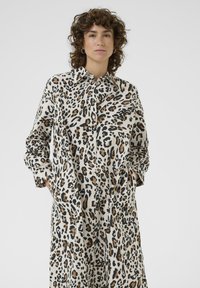 Hnedo-bežová košeľa s leopardím vzorom na gombíky. Má dlhé rukávy, klasický golier a bočné vrecká, vyrobená z ľahkej látky.