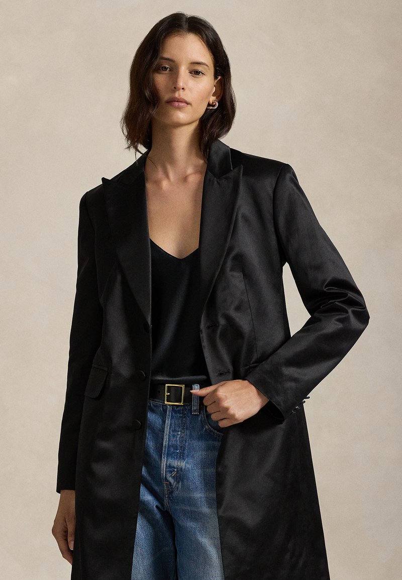 Casaco de cetim preto com corte ajustado, lapelas com entalhe e fechos de botão, combinado com uma blusa preta e jeans azuis desbotados.