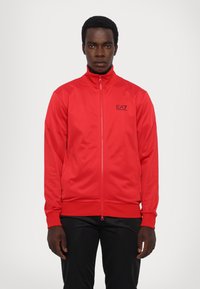 Sarkans jaku zip-up ar augstu apkakli, garām piedurknēm un EA7 logo. Gluda tekstūra, pieguļošs dizains un riboti piedurkņu gali un apakšdaļa.