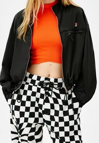 Kvinna som bär en svart zip-jacka över en ljus orange croppad topp, ihop med svart och vit rutiga byxor med dragsko.