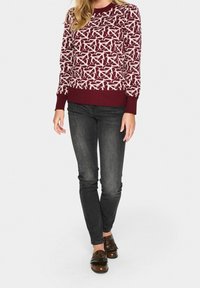 Pull en maille bordeaux avec motif abstrait blanc, col rond, poignets et ourlet côtelés. Associé à un jean foncé et des mocassins marron.