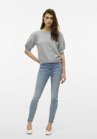 Jasnoszary dzianinowy sweter z bufiastymi rękawami, w zestawie z dopasowanymi niebieskimi dżinsami i beżowymi butami na niskim obcasie. Modelka pozuje z jedną ręką na biodrze.