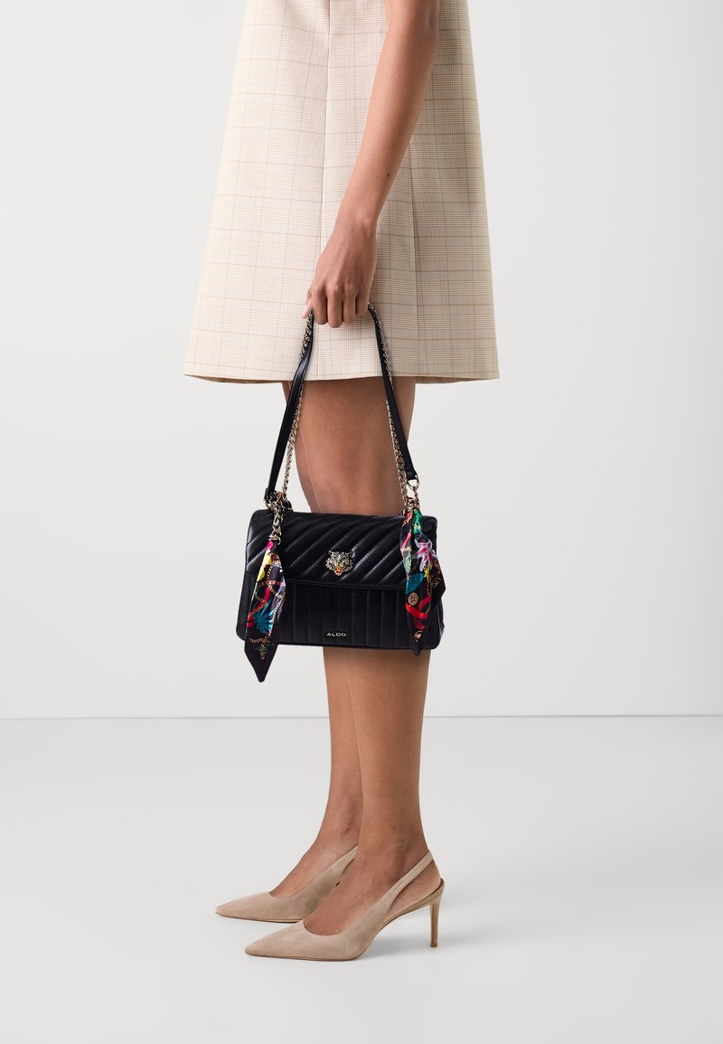 Femme portant des talons beiges et une jupe à carreaux tenant un sac à main noir matelassé avec des foulards colorés attachés aux poignées.