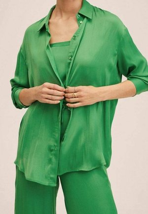 Femme portant une chemise ample verte boutonnée sur un haut et un pantalon verts assortis, en train de fermer les boutons de la chemise avec les deux mains.