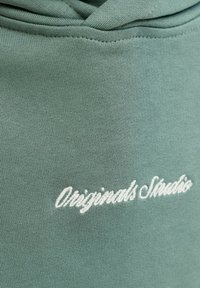 Gros plan sur un tissu vert sauge avec une broderie blanche en écriture cursive portant la mention "Originals Studio" près d’une couture.