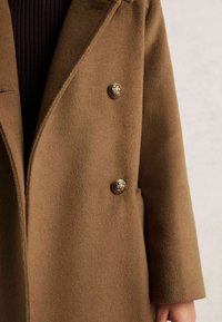 Rudas vilnonis paltas su dekoratyviniais auksiniais mygtukais, dėvimas ant tamsiai juodos raukšlėtos golfos, su tiesiomis rankovėmis ir šonine kišene.