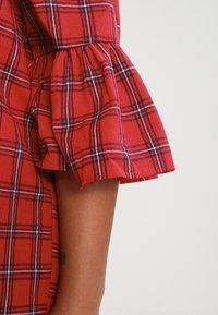 Manche de robe en tartan rouge avec un design froncé et évasé. Le tissu présente un motif à carreaux noir et blanc sur fond rouge.