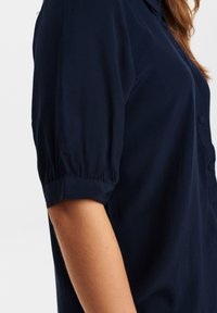 Blusa blu navy con maniche a palloncino corte, in tessuto morbido, chiusura con bottoni frontale e vestibilità sagomata, con una texture liscia e una leggera lucentezza.