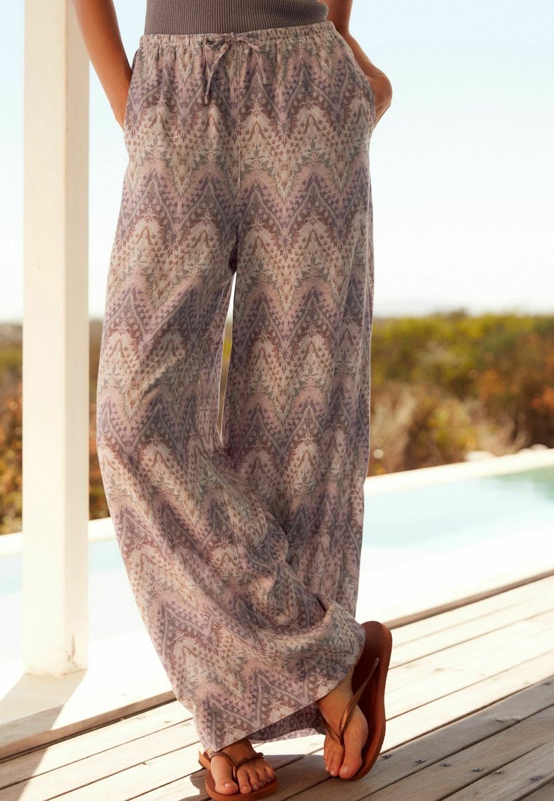 Pantalon ample à motifs avec cordon, porté par une personne debout sur une terrasse en bois près d'une piscine, portant des sandales marron.