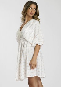 Vestido blanco con rayas doradas, escote en forma de V, mangas holgadas, cintura ajustada y tela texturizada. El dobladillo cae por encima de la rodilla.