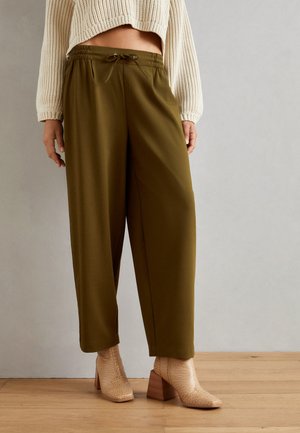Pantalons larges olive, avec une taille élastique et un cordon de serrage, associés à des bottes à talons texturées beige. Fond en bois.