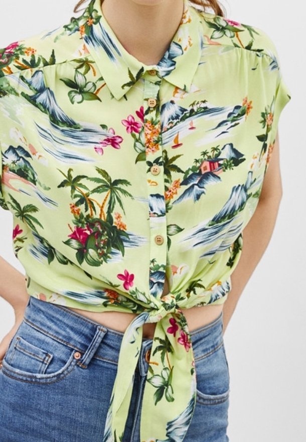 Blommig kortärmad skjorta i ljusgrön med knäppning framtill, prydd med mönster av berg, palmer och blommor, knuten i midjan.