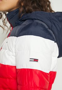 Veste rembourrée colorée en rouge, blanc et marine avec capuche, dotée d'un patch logo sur la manche et d'une texture matelassée. Zipper visible.