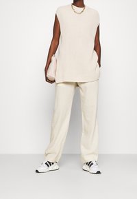 Bezruký béžový pletený top s vysokým límcem, v kombinaci s krémovými širokými kalhotami a bílými teniskami s černými akcenty. Drží malou béžovou tašku.