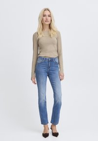 Blonde Frau steht in einem beigen Langarmshirt, blauen Jeans und schwarzen spitzen Schuhen vor einem schlichten hellen Hintergrund.