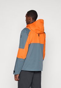 Άνδρας με πλάτη που φοράει μια μπλε και πορτοκαλί φούτερ με κουκούλα και μακριά μανίκια, συνδυασμένο με σκούρο παντελόνι, ενάντια σε απλό φόντο.