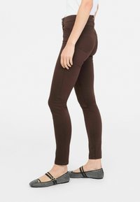 Jeans skinny marroni realizzati in tessuto elasticizzato, dotati di vita alta e tasche posteriori, abbinati a ballerine con motivo in bianco e nero e cinturini.