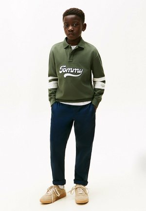 Pull vert avec des rayures blanches, arborant le logo "Tommy". Un pantalon bleu marine et des baskets beiges complètent la tenue, mettant en avant un style décontracté.