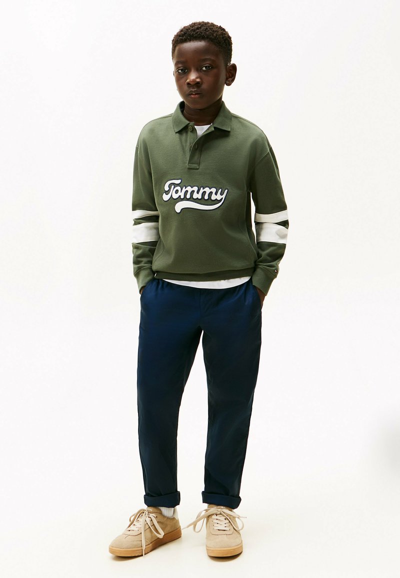 Groene trui met witte strepen, met het 'Tommy' logo. Marineblauwe broek en beige sneakers maken de outfit compleet, wat een casual stijl laat zien.