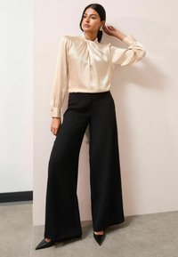 Spīdīga, gaiši zelta garām piedurknēm blūze ar plecu detalizāciju, apvienota ar platām melnā krāsā biksēm un norādītām melnajām kurpēm.