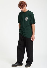 Grøn bomulds-t-shirt med et broderet logo på forsiden, parret med sorte brede bukser og sorte snørebåndssko, sat mod en ensfarvet baggrund.