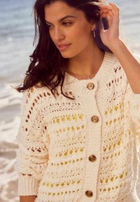 Cardigan en maille crème avec motif ajouré, doté de cinq boutons en bois et d'une coupe décontractée. Une légère texture améliore son design.