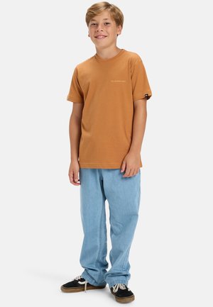 Camiseta de algodón marrón de manga corta con un pequeño logo, combinada con unos vaqueros sueltos de color azul claro y zapatillas negras.
