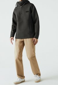 Sweat à capuche gris foncé avec poche avant et fermeture éclair au quart. Pantalon beige, baskets blanches avec une semelle claire, et un logo discret sur la poitrine.