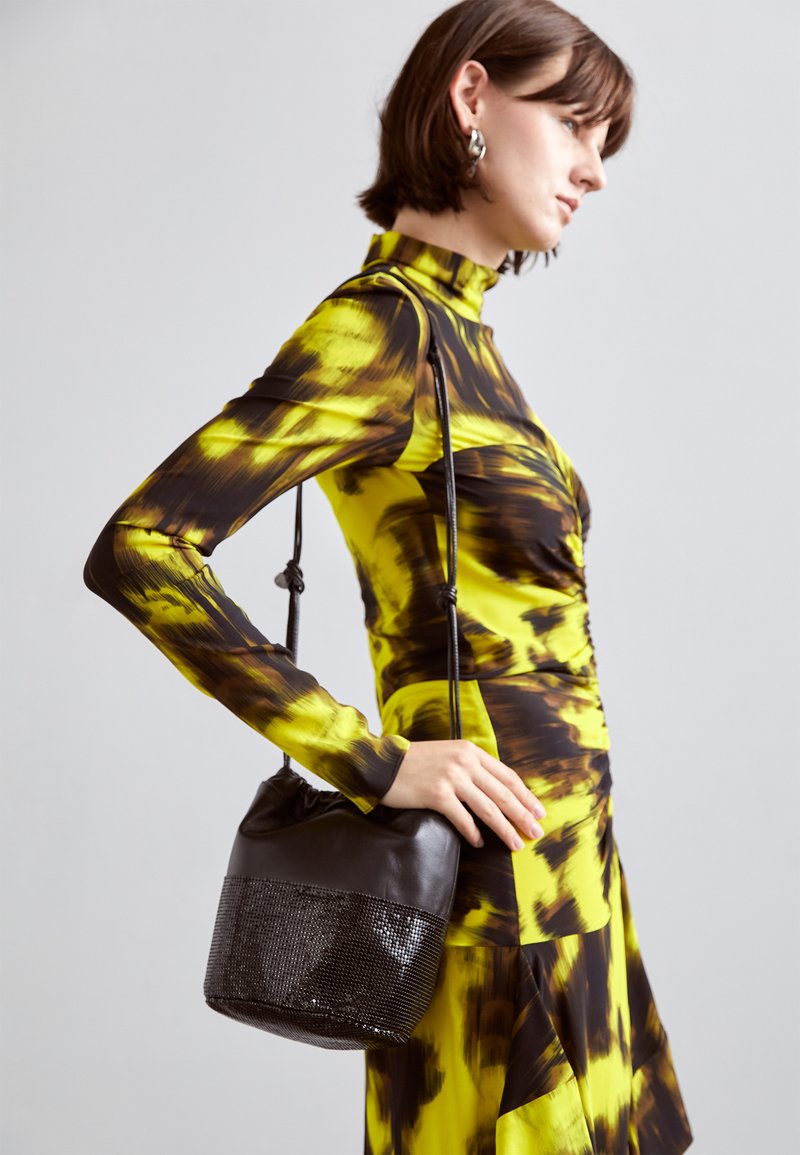 Sac din piele neagră tip găleată, având o bază din plasă, cu o textură moale și o curea subțire pentru umăr. Este purtat împreună cu o rochie cu model galben și maro.