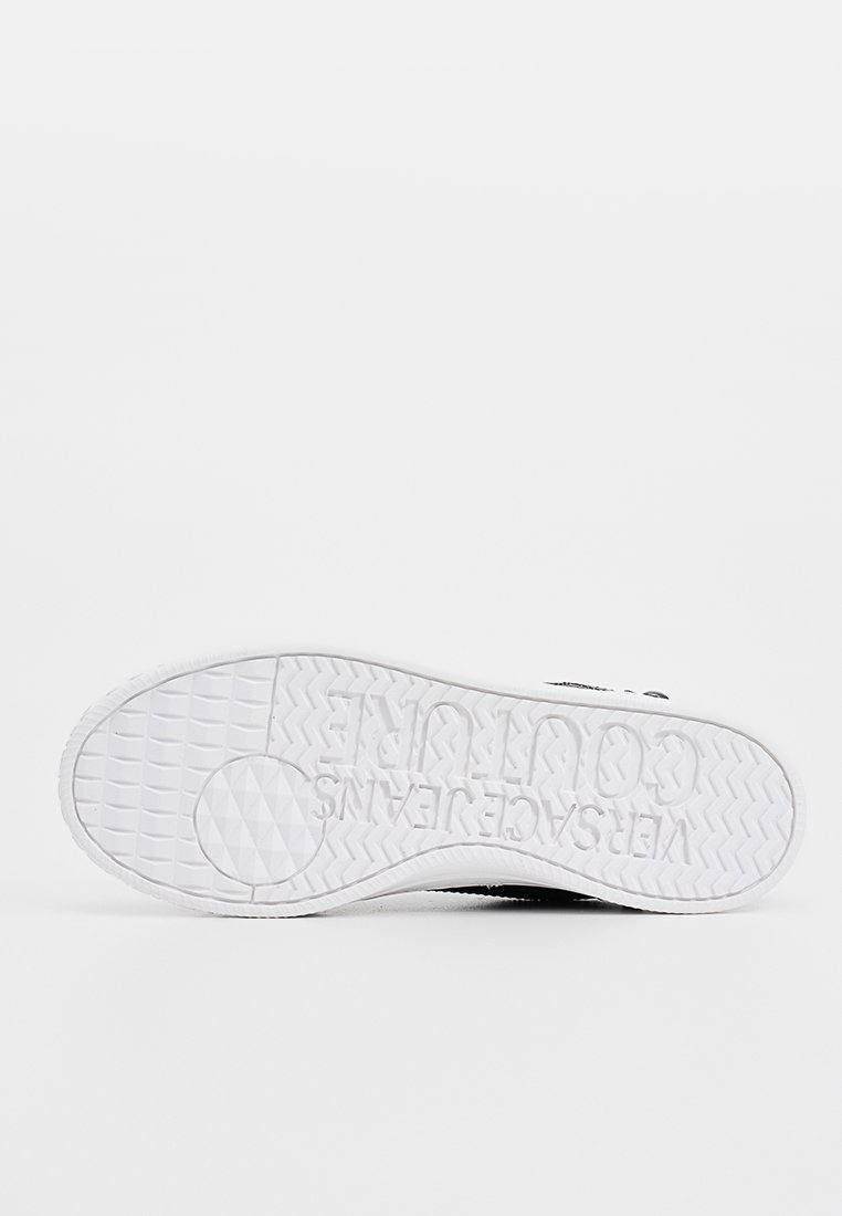 Suela de una zapatilla blanca con agarre texturizado y el logo "WERSACE JEANS COUTURE" en relieve en reversa.