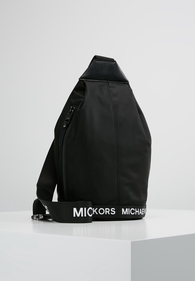 Czarna torba na ramię Michael Kors z kieszenią na zamek i regulowanym paskiem z logo, prezentowana na białym piedestale na jednolitym tle.