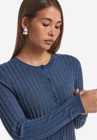 Niebieski kardigan z żebrowanym wykończeniem i okrągłym dekoltem, z sześcioma guzikami i długimi rękawami. Tkanina ma teksturowany wygląd, prezentując wzory w warkocz.